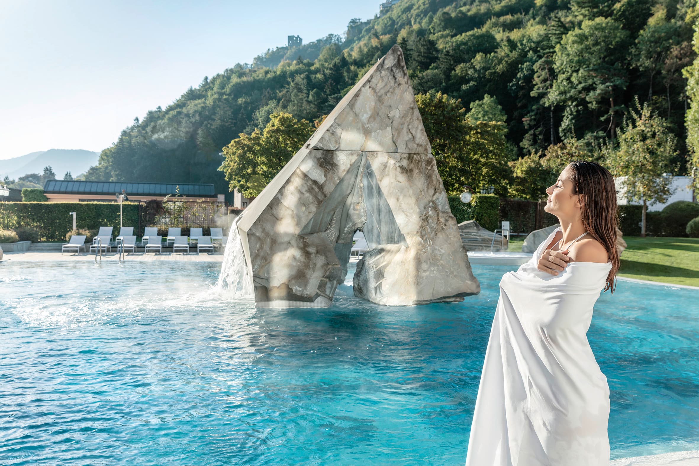 Eine Frau eingehüllt in einem Badetuch steht vor der Außenanlage der Tamina Therme und hat das Gesicht zur Sonne geneigt.