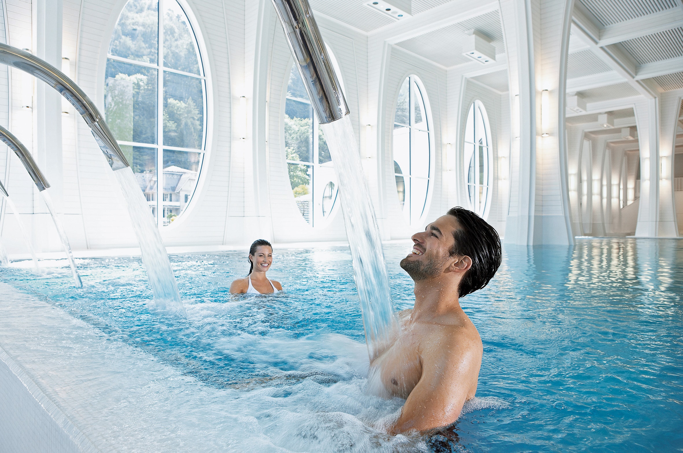 Ein Mann steht unter einer Nackendusche und im Hintergrund schwimmt eine Frau im Thermalbad der Tamina Therme.