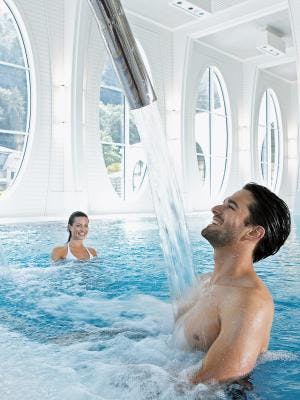 Ein Mann steht unter einer Nackendusche und im Hintergrund schwimmt eine Frau im Thermalbad der Tamina Therme